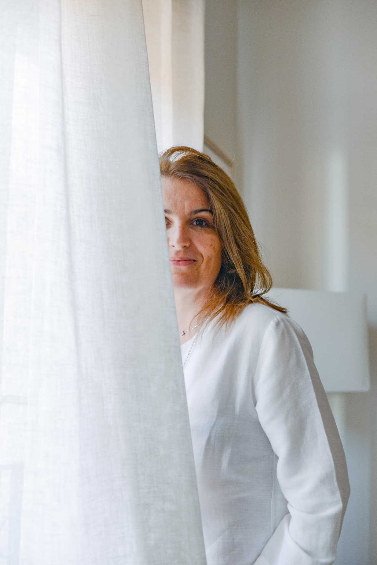 Fah Maioli looking out from behind a white linen curtain in soft daylight